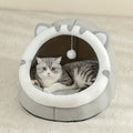 Grey and white cat-ear cave with a round opening and a hanging pom toy, a tabby cat lying inside