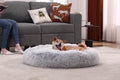 Grey round faux-fur donut dog bed on a rug with a shiba lying in it, beside a sofa