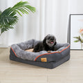 Dark grey rectangular bolster dog bed with orange piping and a beige plush centre, a small black dog lying in it