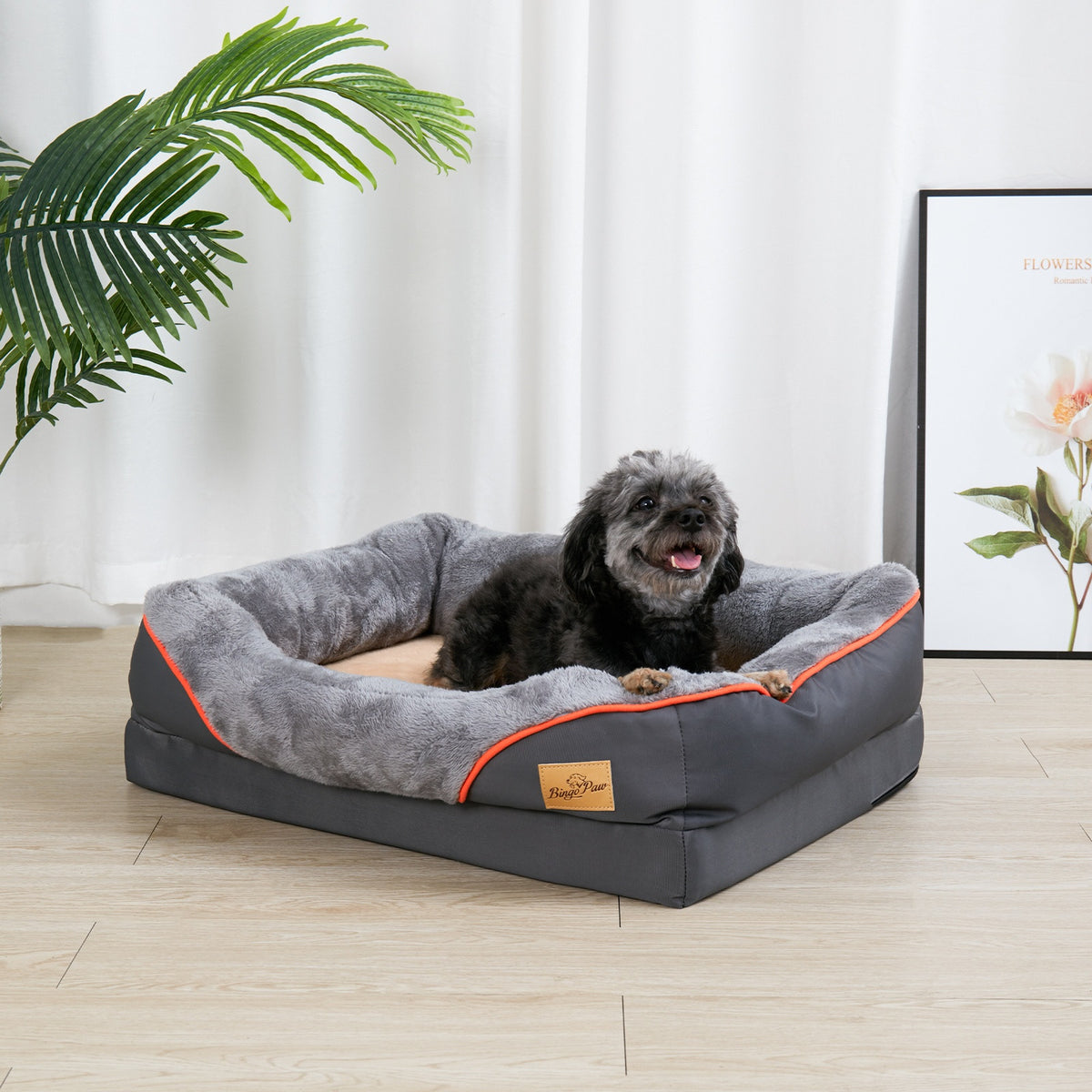 Dark grey rectangular bolster dog bed with orange piping and a beige plush centre, a small black dog lying in it