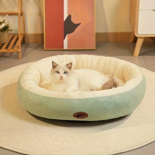 Sage green oval pet bed with a cream plush lining and a round brown pictogram patch, a white cat lying in it