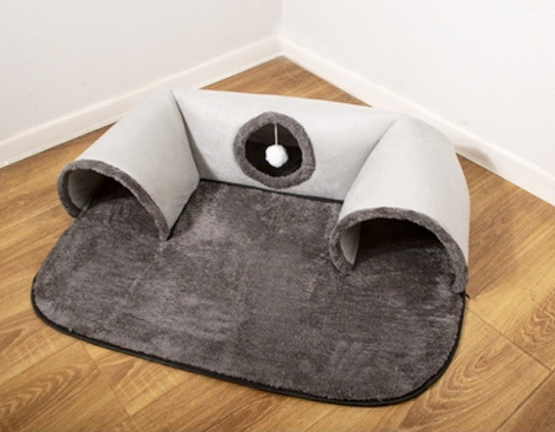 Grey U-shaped plush cat tunnel on a flat dark grey plush mat, in a room corner