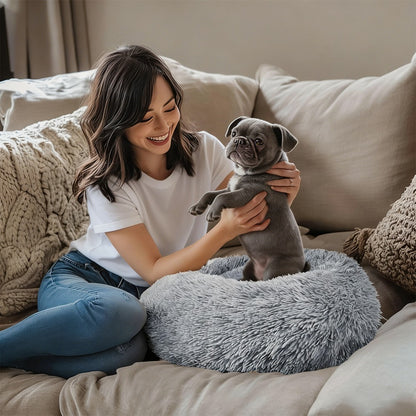 A woman on a sofa holds a grey puppy sitting in the grey shag donut bed