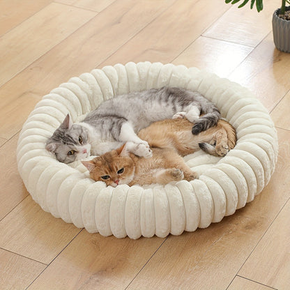 A grey tabby and a ginger cat lie together in the round cream plush bed on a wooden floor