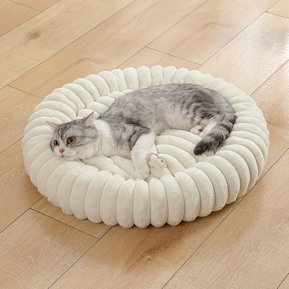 A grey tabby cat stretches out in the round cream plush bed on a wooden floor