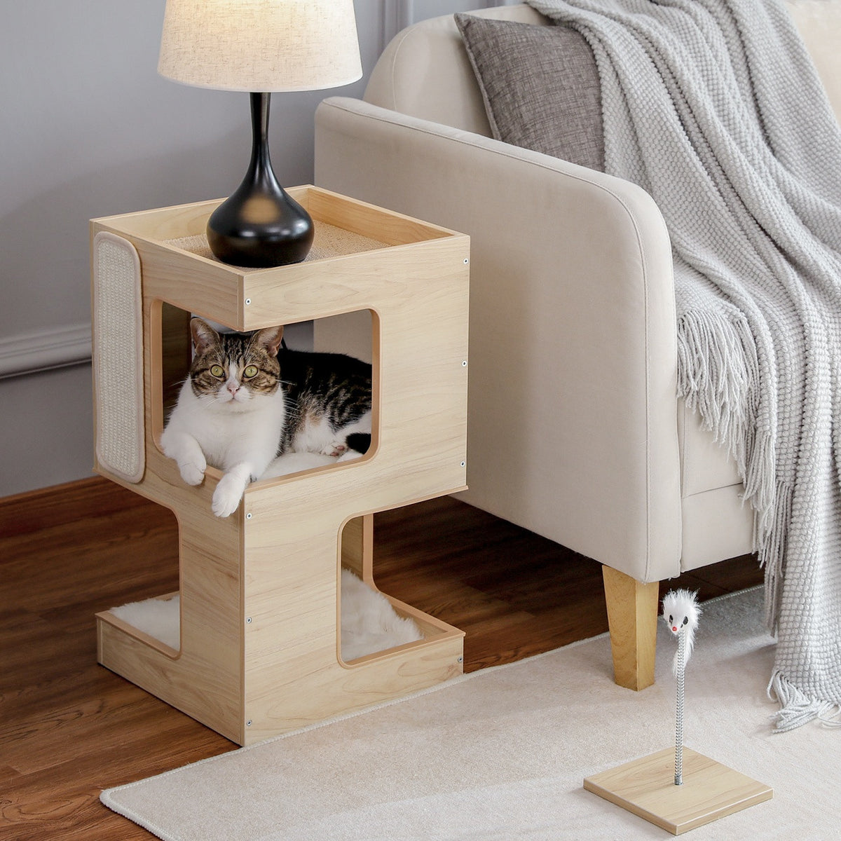 Natural wood cat condo beside a sofa with a cat resting on the upper level and the spring mouse toy on the rug